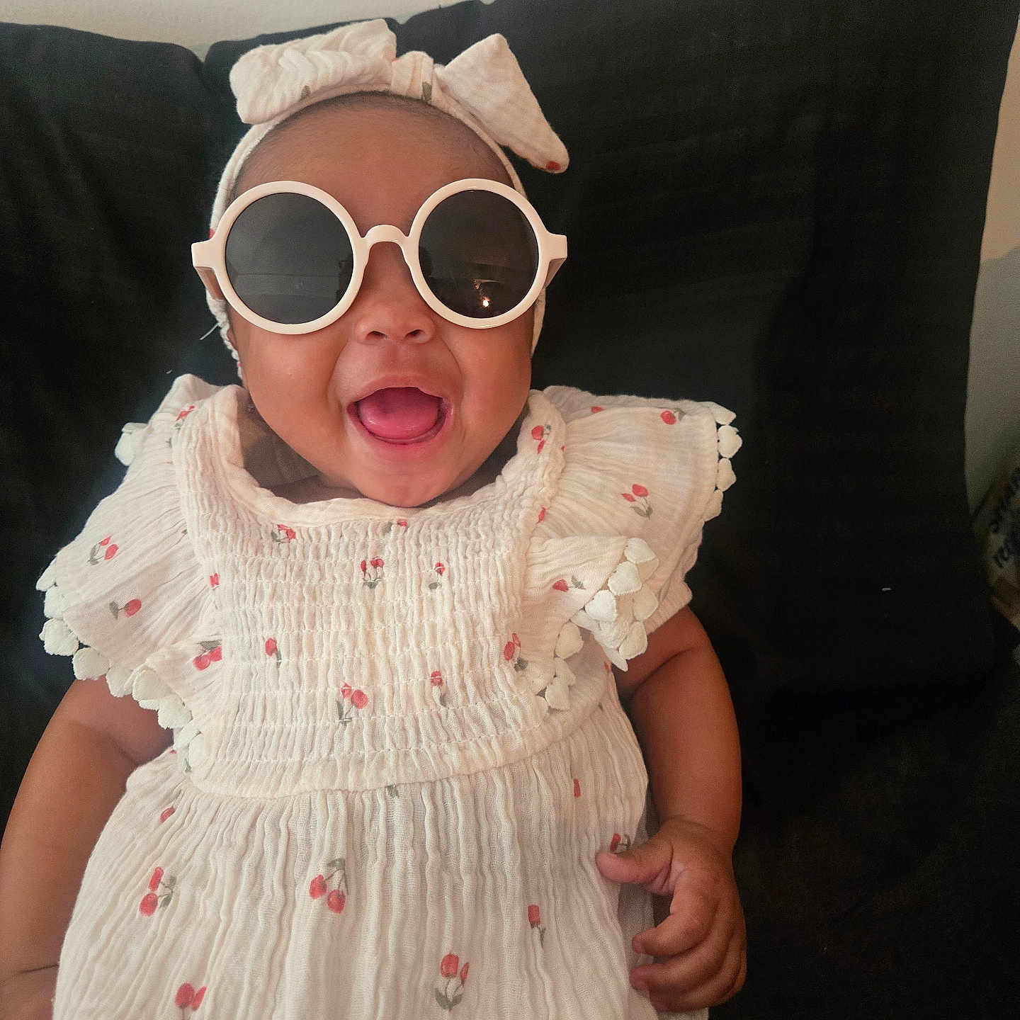 Azure is registered to the contest to win money with this photo: baby, child, sunglasses, headband, bow, white_dress, floral_pattern, happy, smiling, indoors, portrait, cute, infant, sitting, fashion, accessory, dark_background, cheerful, person, closeup