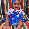 baby, blue_bow, celebration, child, clothing, colorful, cute, face, fashion, feet, happy, headband, infant, outdoor, person, portrait, red_stars, sitting, smiling, striped_cushion