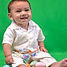 baby, child, smiling, sitting, green_background, easter_eggs, toy_carrots, white_shirt, floral_pants, barefoot, cute, portrait, studio_photo, infant, happy, colorful, playful, indoors, one_person, toddler