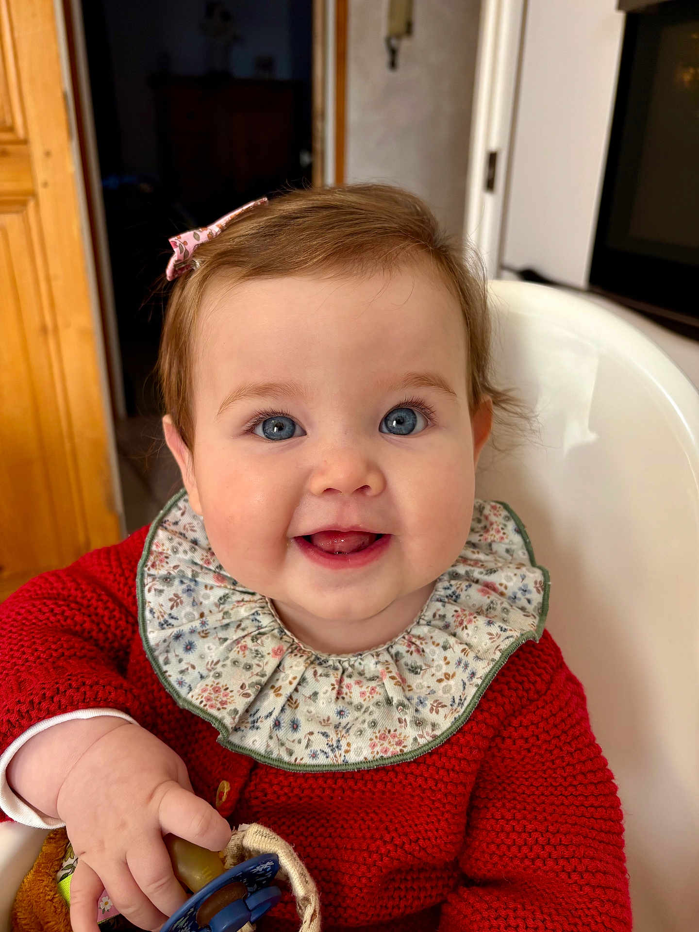 Victoire participe au concours pour gagner de l'argent avec cette photo : baby, child, smiling, blue_eyes, red_sweater, floral_collar, high_chair, indoors, cute, happy, face, hand, pacifier, person, infant, portrait, seated, clothing, furniture, warm_lighting
