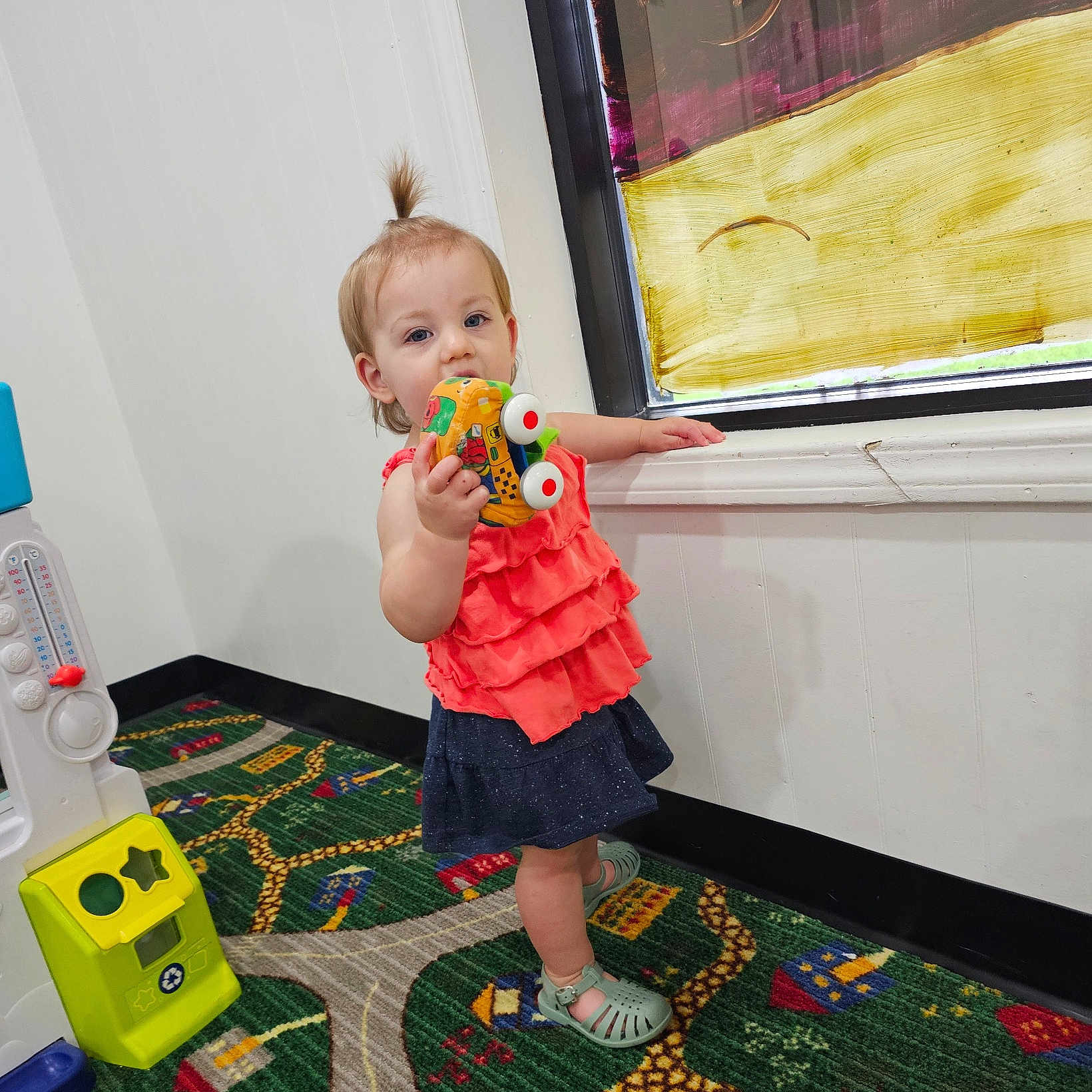 Aurora is registered to the contest to win money with this photo: carpet, child, clothing, colorful, curtain, footwear, holding, indoor, patterned_carpet, person, playful, playroom, sandal, standing, toddler, topknot_hairstyle, toy_car, wall, window, young_child