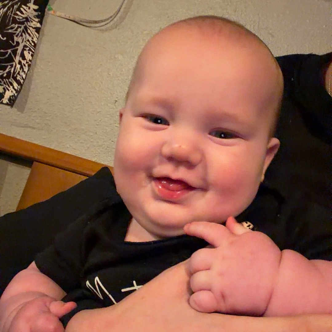 Onyx is registered to the contest to win money with this photo: baby, smiling, infant, chubby_cheeks, person, hand, holding, indoor, wall, curtain, black_clothing, happy, cute, young_child, portrait, skin, face, adult_hand, seated, domestic