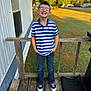 child, boy, smiling, glasses, striped_shirt, polo_shirt, jeans, sneakers, porch, wooden_deck, railing, mobile_home, lawn, grass, trees, outdoor, sunlight, portrait, hands_in_pockets, casual