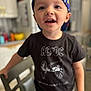 child, toddler, boy, baseball_cap, cap_backwards, acdc_shirt, t_shirt, smiling, teeth, eyes, face, kitchen, chair, countertop, indoor, portrait, candid, standing, shallow_depth_of_field, casual_clothing