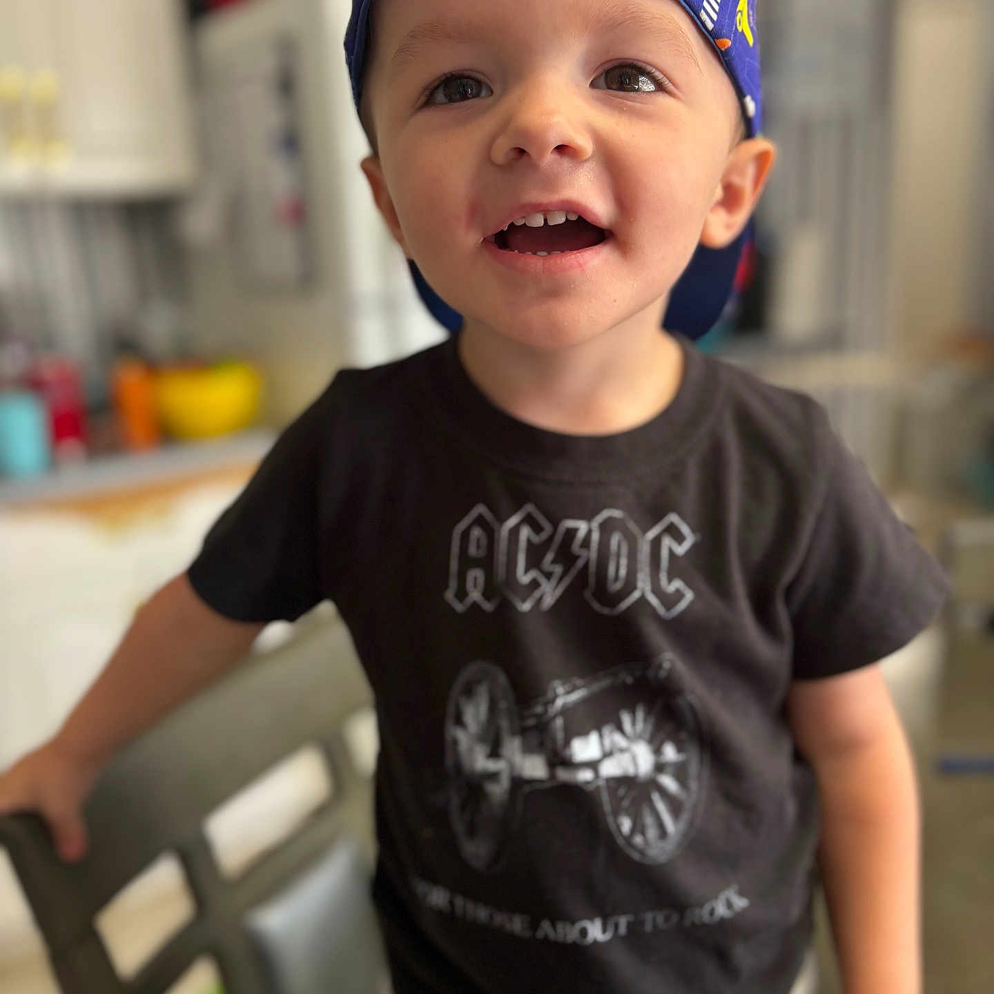 Onyx is registered to the contest to win money with this photo: acdc_shirt, baseball_cap, boy, candid, cap_backwards, casual_clothing, chair, child, countertop, eyes, face, indoor, kitchen, portrait, shallow_depth_of_field, smiling, standing, t_shirt, teeth, toddler