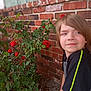 child, boy, red_roses, brick_wall, green_leaves, outdoor, garden, flower, plant, nature, casual_clothing, short_sleeves, young, portrait, side_view, daylight, shrubbery, ground, relaxed, natural_light