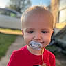 child, toddler, baby, portrait, outdoor, sunlight, red_shirt, pacifier, blond_hair, blue_eyes, smiling, face, close_up, daylight, backyard, grass, sidewalk, house, vehicle, cord