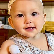Esther is registered to the contest to win money with this photo: baby, child, headband, flower, plush_toy, bunny, dress, patterned_clothing, hand, indoor, home, furniture, shelf, picture_frame, smiling, cute, portrait, person, infant, soft_toy
