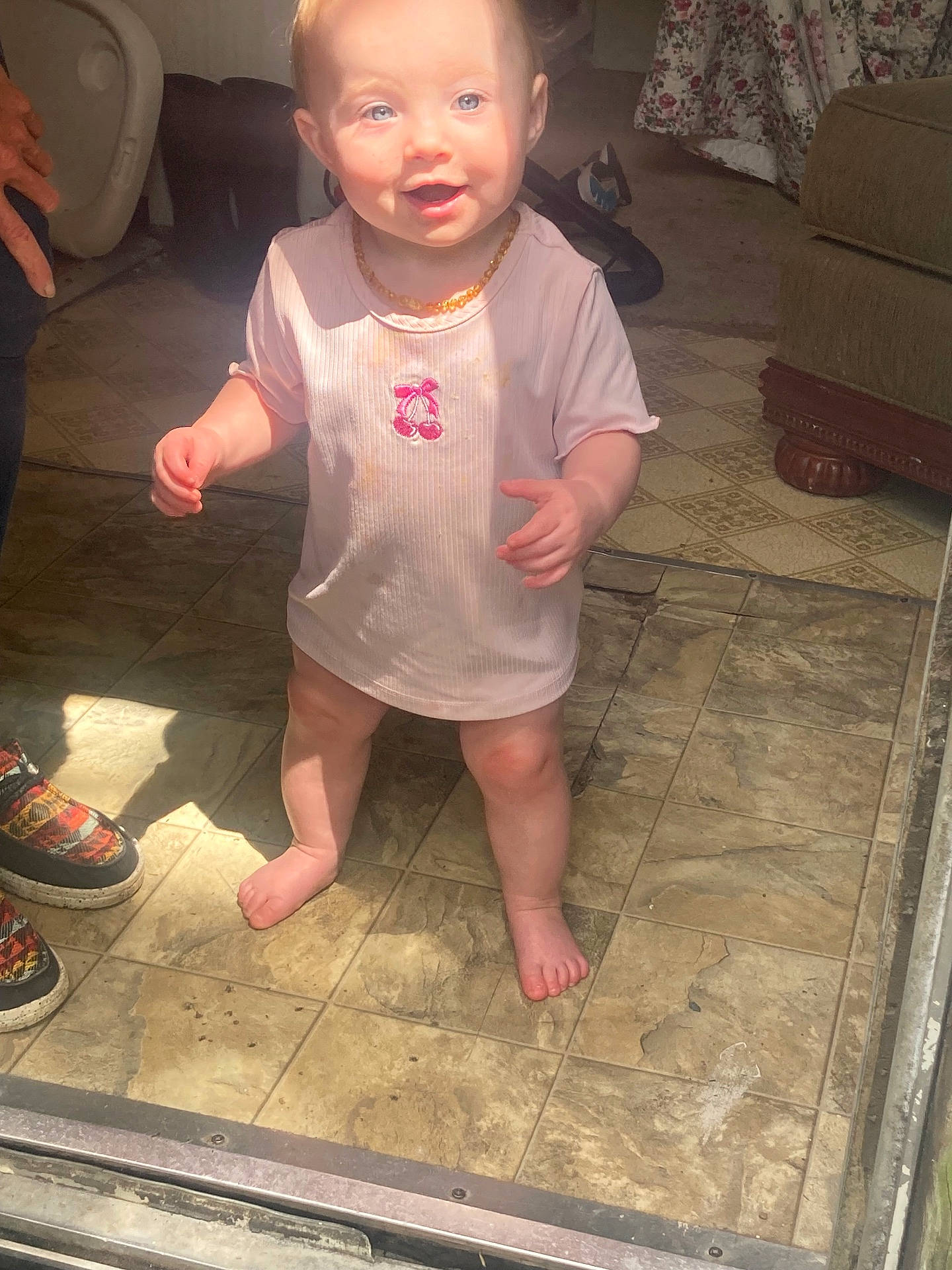 Kodi is registered to the contest to win money with this photo: baby, barefoot, child, curtain, face, hands, happy, home, indoor, living_room, necklace, portrait, shirt, shoes, smiling, sofa, standing, sunlight, tile_floor, toddler