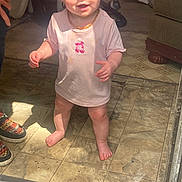 Kodi is registered to the contest to win money with this photo: baby, child, toddler, smiling, standing, portrait, indoor, living_room, tile_floor, shirt, necklace, barefoot, face, hands, sunlight, curtain, sofa, shoes, home, happy
