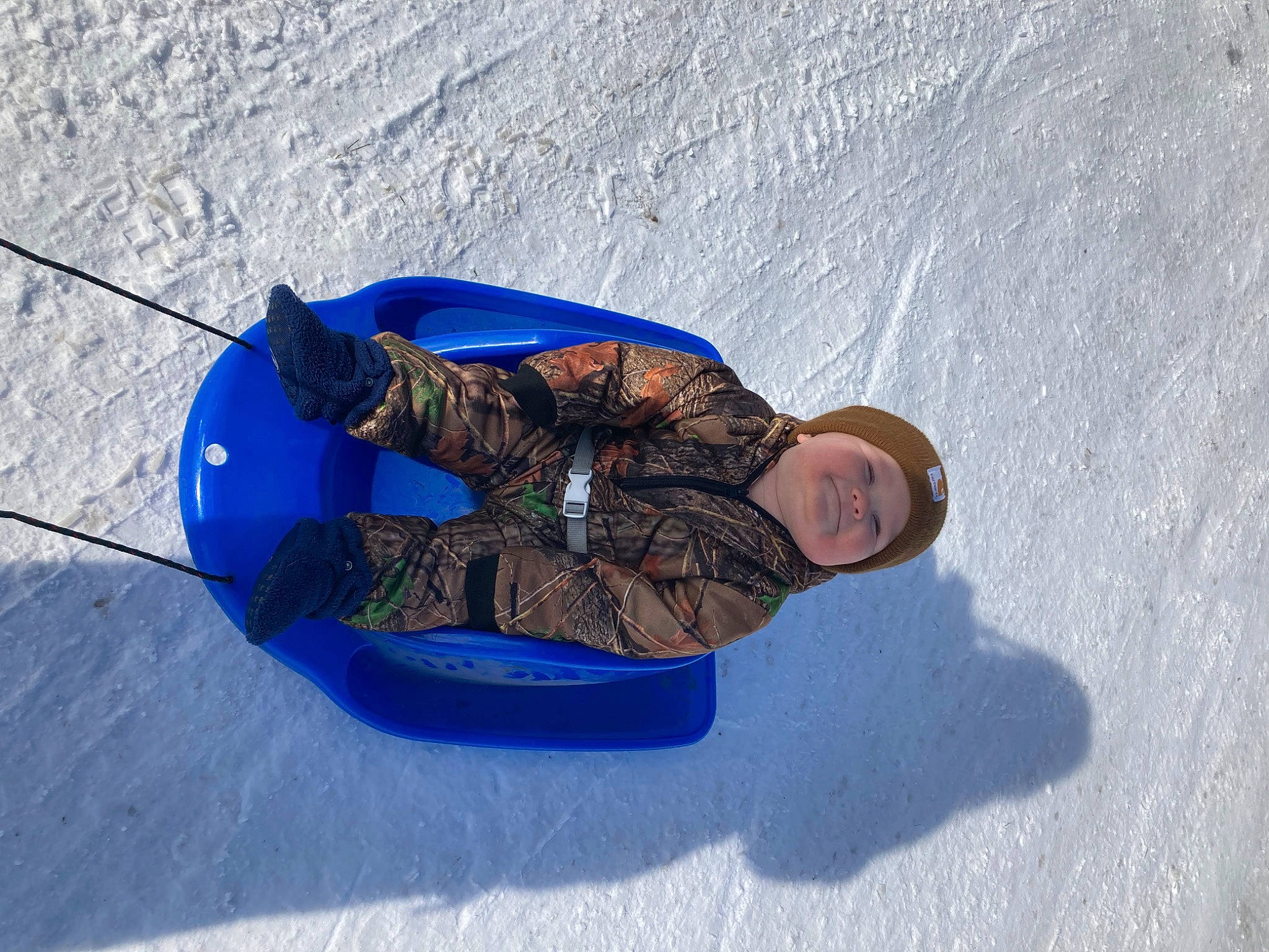 Briggs is registered to the contest to win money with this photo: boats_and_boating_equipment_and_supplies, concrete, electric_blue, freezing, fun, headwear, individual_sports, jacket, joy, leisure, person, recreation, shadow, sitting, sled, sledding, slope, smile, snow, sports