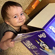 Elijah is registered to the contest to win money with this photo: toddler, child, baby, carpet, box, cartoon, smile, face, hand, indoor, young_child, clothing, cute, person, playful, expression, floor, toy_box, short_hair, happy