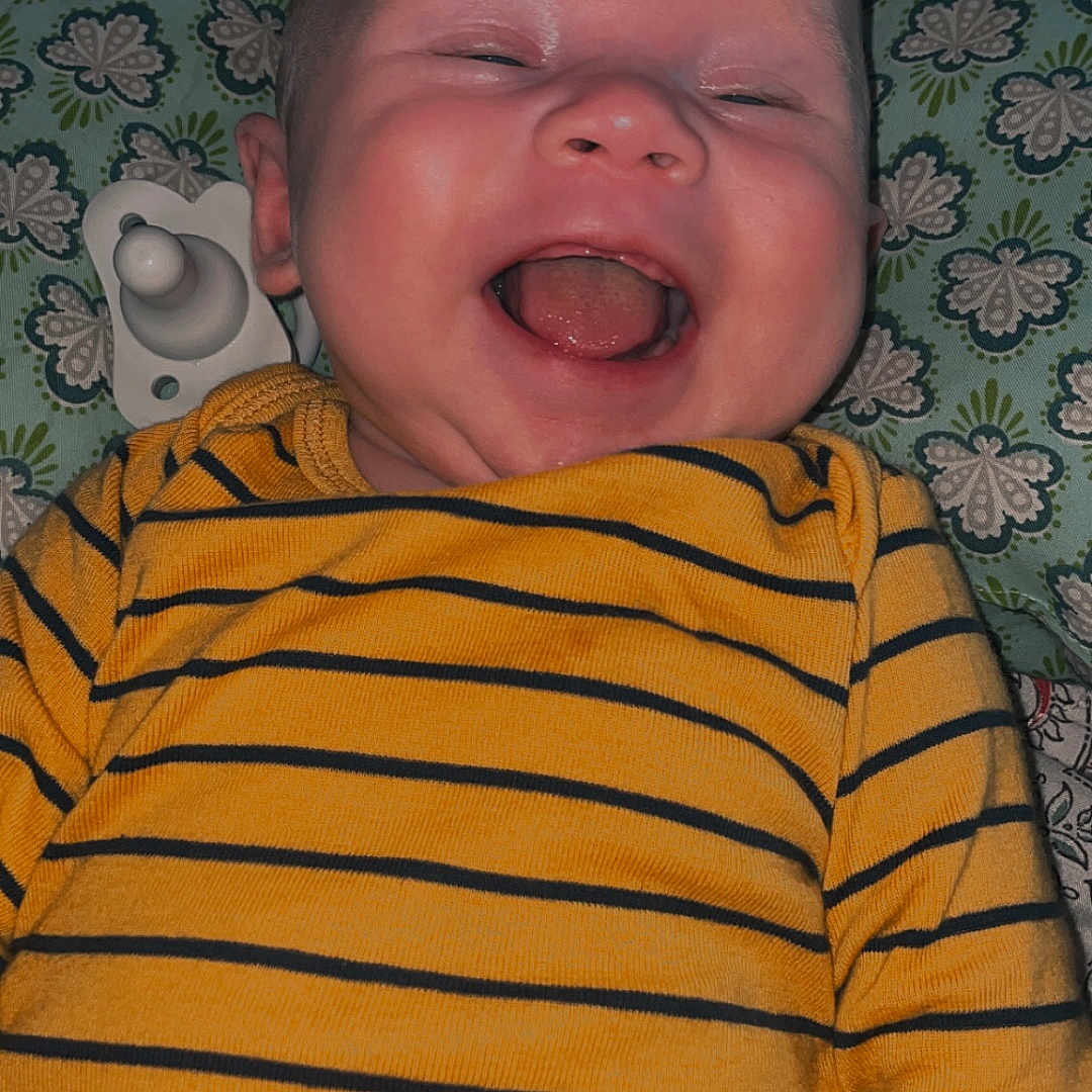 Alijha is registered to the contest to win money with this photo: baby, child, close_up, cushion, cute, face, happy, head, indoors, infant, laughing, lying_down, mouth_open, pacifier, patterned_background, person, portrait, smiling, striped_clothing, yellow_shirt