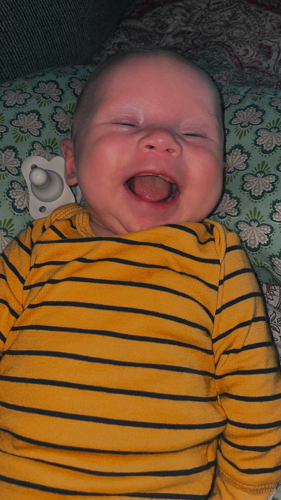 Alijha is registered to the contest to win money with this photo: baby, infant, smiling, laughing, yellow_shirt, striped_clothing, pacifier, cushion, patterned_background, happy, child, face, mouth_open, lying_down, close_up, cute, indoors, portrait, person, head