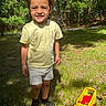 child, smiling, outdoor, grass, trees, toy, yellow_toy, shorts, tshirt, sunlight, nature, happy, play, boy, forest, daylight, feet, shadow, casual_clothing, park