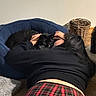 Millie is registered to the contest to win money with this photo: cat, person, indoor, pet_bed, black_cat, black_clothing, plaid_pants, carpet, cuddling, affection, glasses, basket, wall, floor, cozy, resting, human_pet_interaction, long_hair, home, relaxation