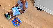 Seyven is registered to the contest to win money with this photo: child, toddler, smile, tablet, floor, food, snack, plate, socks, hair, doorway, indoors, wooden_floor, technology, happy, casual, playing, person, clothing, fun