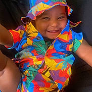 Seyven is registered to the contest to win money with this photo: toddler, child, smiling, colorful_clothing, hat, bucket_hat, happy, portrait, baby, casual, cute, indoor, person, fashion, playful, fun, bright_colors, sitting, young_child, joyful