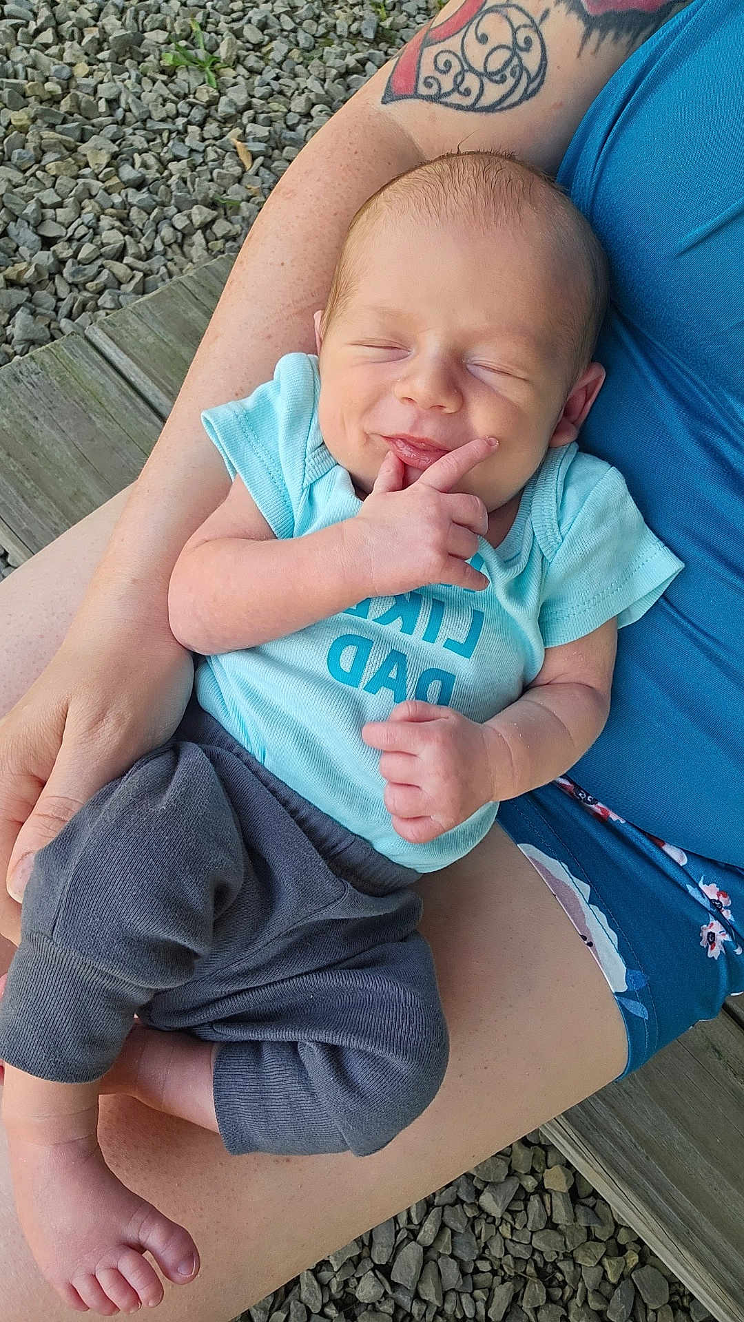 Nash joined the competition — help win amazing prizes! baby, infant, sleeping, person, tattoo, blue_clothing, gray_pants, hand, foot, outdoor, gravel, wooden_planks, skin, arm, lap, closeup, cute, peaceful, casual, relaxing