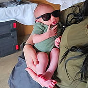 Nash is registered to the contest to win money with this photo: baby, infant, sunglasses, green_onesie, person, holding, arm, hand, indoor, table, stroller, foot, skin, casual, relaxed, cool, tiny, adult, background, blur