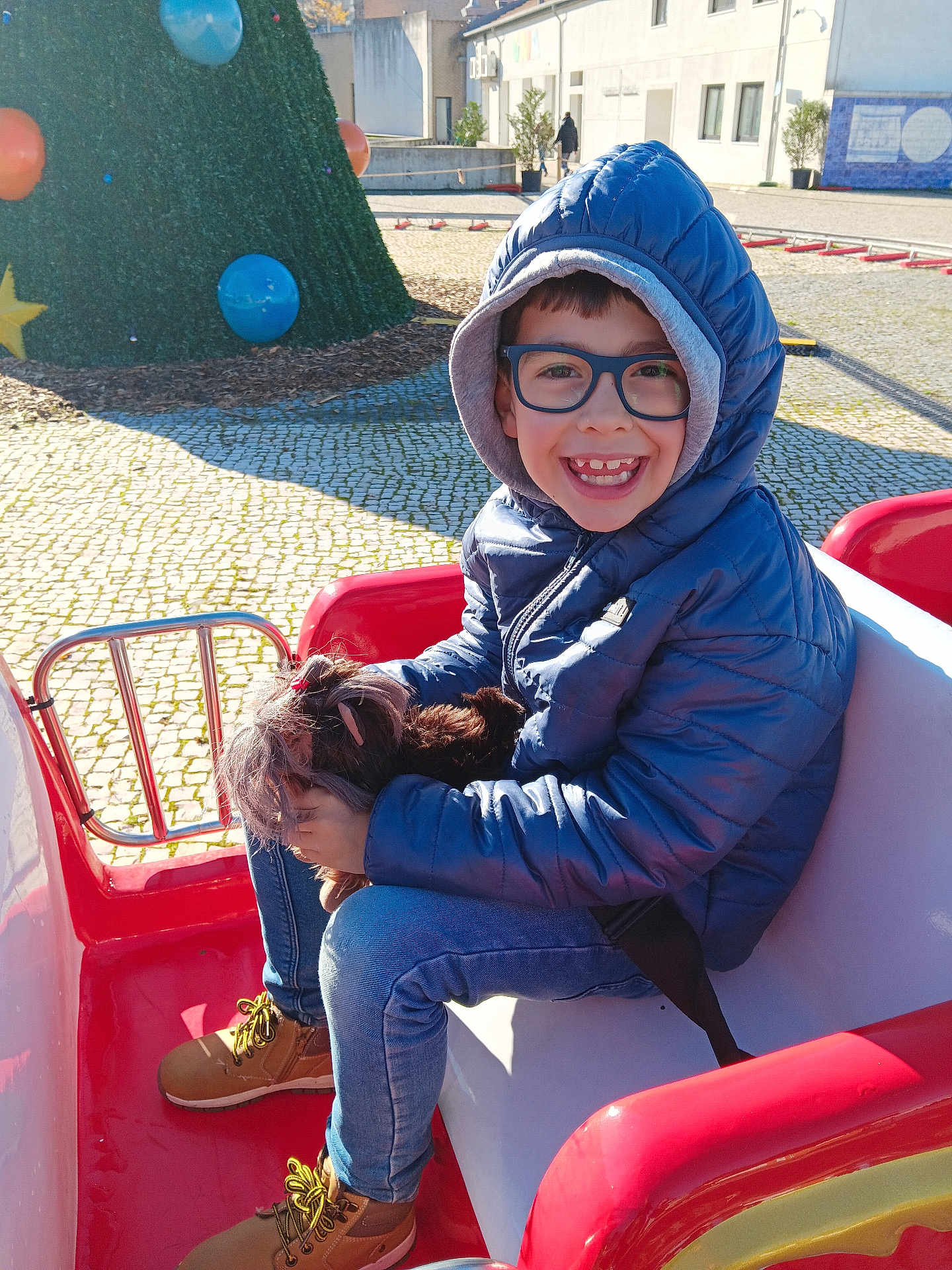 Claudio Teixeira participe au concours pour gagner de l'argent avec cette photo : child, boy, glasses, blue_jacket, hood, smiling, dog, pet, red_ride, outdoor, sunny, balloons, christmas_tree, pavement, casual_clothing, jeans, boots, happy, playground, daytime