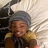 child, smiling, peace_sign, blanket, head_wrap, bed, pillow, yellow_shirt, happy, indoor, portrait, person, young_child, cozy, casual, comfort, relaxed, home, cute, gesture