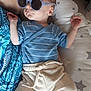 baby, infant, sunglasses, blue_clothing, striped_shirt, beige_pants, socks, text_on_socks, star_pattern, cushion, arm, relaxing, indoor, person, cute, sleepy, small_hands, headwear, eyewear, comfort