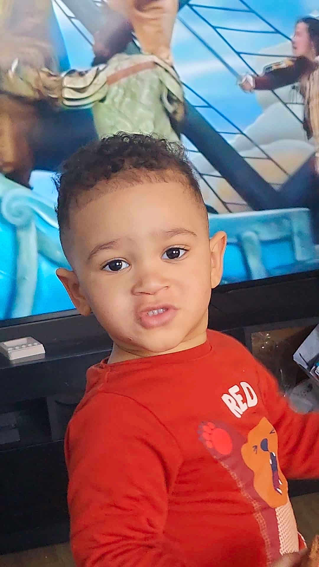 Zayn participe au concours pour gagner de l'argent avec cette photo : child, toddler, boy, portrait, face, curly_hair, big_eyes, red_shirt, lion_graphic, expression, living_room, television, tv_show, background_characters, ear, smile, close_up, furniture, remote_control, floor