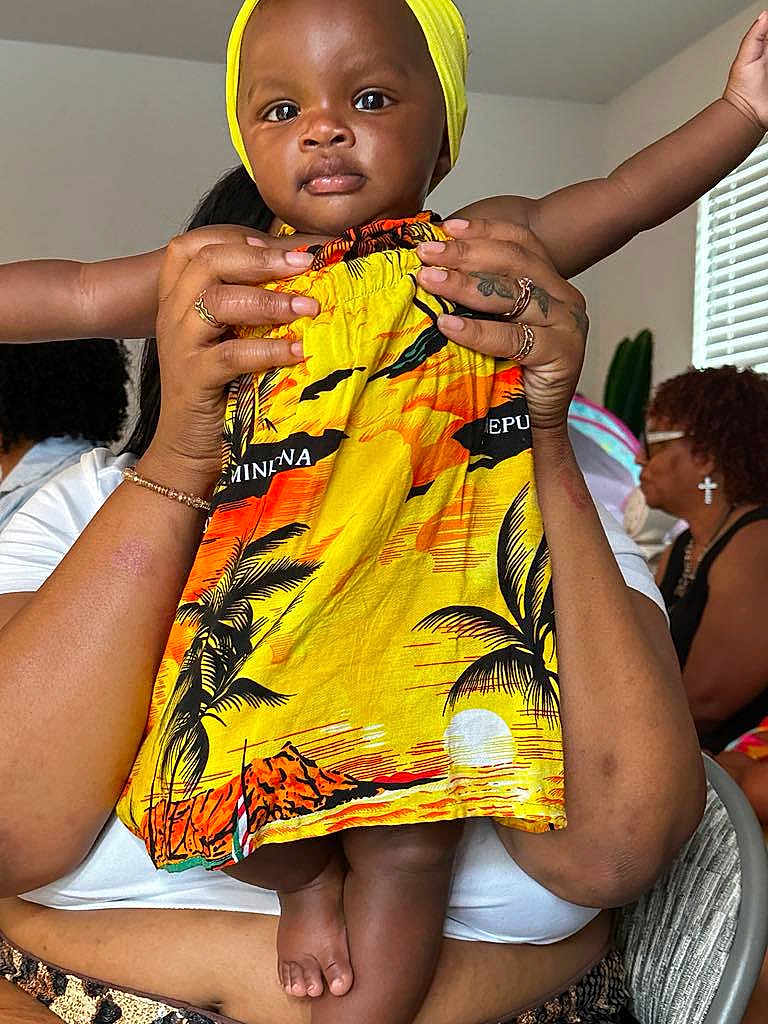 Mulani is registered to the contest to win money with this photo: baby, child, toddler, face, hands, feet, dress, yellow, orange, patterned_clothing, adult_hands, indoor, window, people, seated, room, casual, portrait, holding, family