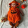baby, pumpkin_costume, hooded_outfit, striped_leggings, blanket, knitted_pumpkin, orange, white, flowers, smiling, cute, infant, lying_down, fall_theme, decor, cozy, child, portrait, festive, holiday