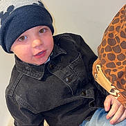 Flavio participe au concours pour gagner de l'argent avec cette photo : black_jacket, blanket, blue_jeans, boy, casual_clothing, child, closeup, curious, face, giraffe_pattern, hat, indoor, jacket, jeans, knit_hat, light_background, looking_up, person, seated, warm_clothing