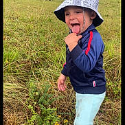 Flavio participe au concours pour gagner de l'argent avec cette photo : bracelet, bucket_hat, child, cow, cows, daytime, farmhouse, field, grass, jacket, outdoors, pants, pasture, playful, portrait, rural, screenshot, smiling, toddler, tongue_out
