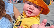 Ethan is registered to the contest to win money with this photo: adult_hand, baby, blue_dress, building, child, crowd, cute, daylight, face, hat, holding, orange_hat, outdoor, person, reaching_hand, smiling, street, summer, sunny, yellow_shirt