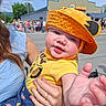 Ethan is registered to the contest to win money with this photo: baby, child, hat, orange_hat, yellow_shirt, outdoor, daylight, person, smiling, reaching_hand, adult_hand, crowd, building, street, blue_dress, summer, sunny, face, cute, holding
