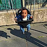 toddler, child, swing, playground, denim_jacket, jeans, cap, smile, outdoor, sunlight, shadow, rubber_matting, metal_fence, green_door, urban, happy, baby_swing, person, clothing, play