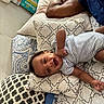 adult_arm, baby, bed, child, cushion, face, hand, happy, home, indoor, infant, lying_down, patterned_textile, person, pillow, resting, skin, sleepwear, smiling, striped_clothing