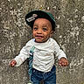 toddler, child, smiling, cap, baseball_cap, barcode, white_sweatshirt, denim_jeans, pacifier, clip, standing, wall, textured_wall, outdoor, happy, person, clothing, casual, cute, portrait