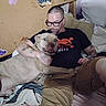 man, person, dog, large_dog, pet, owner, human_animal_bond, cuddling, sitting, lying_down, bed, bedroom, pillows, blanket, clothing, glasses, tattoos, black_shirt, indoor, portrait