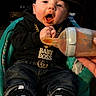 baby, infant, child, portrait, sitting, baseball_cap, black_clothing, gold_chain, necklace, baby_bottle, feeding, food, mouth, hands, sneakers, jeans, high_chair, indoor, looking_at_camera, funny
