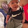 baby, child, infant, portrait, smiling, wide_eyed, bunny_hat, hat, black_shirt, sportswear, swoosh_logo, gray_pants, teddy_bear_booties, shoes, sitting, indoor, bed, blanket, adult, cozy