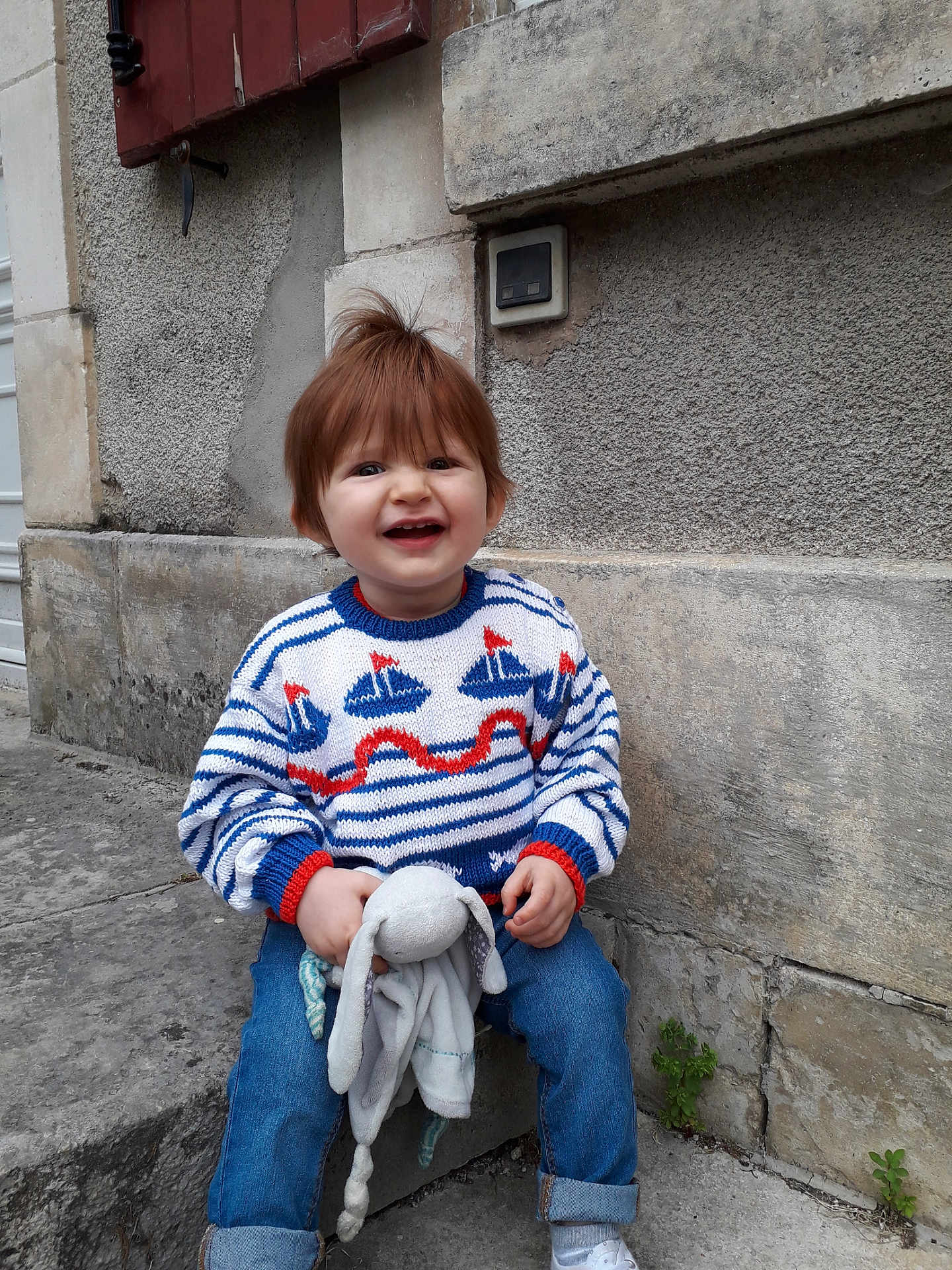 Robin participe au concours pour gagner de l'argent avec cette photo : toddler, child, smiling, sweater, toy, stuffed_animal, jeans, white_shoes, outdoor, stone_step, wall, red_hair, cute, happy, casual_clothing, playful, young_child, sitting, daylight, urban