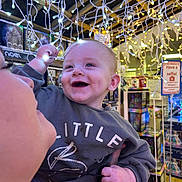 Reggie is registered to the contest to win money with this photo: baby, child, smiling, lights, string_lights, dimples, sweater, person, hand, store, holiday, festive, decoration, indoor, happy, face, holding, adult, cheerful, shopping