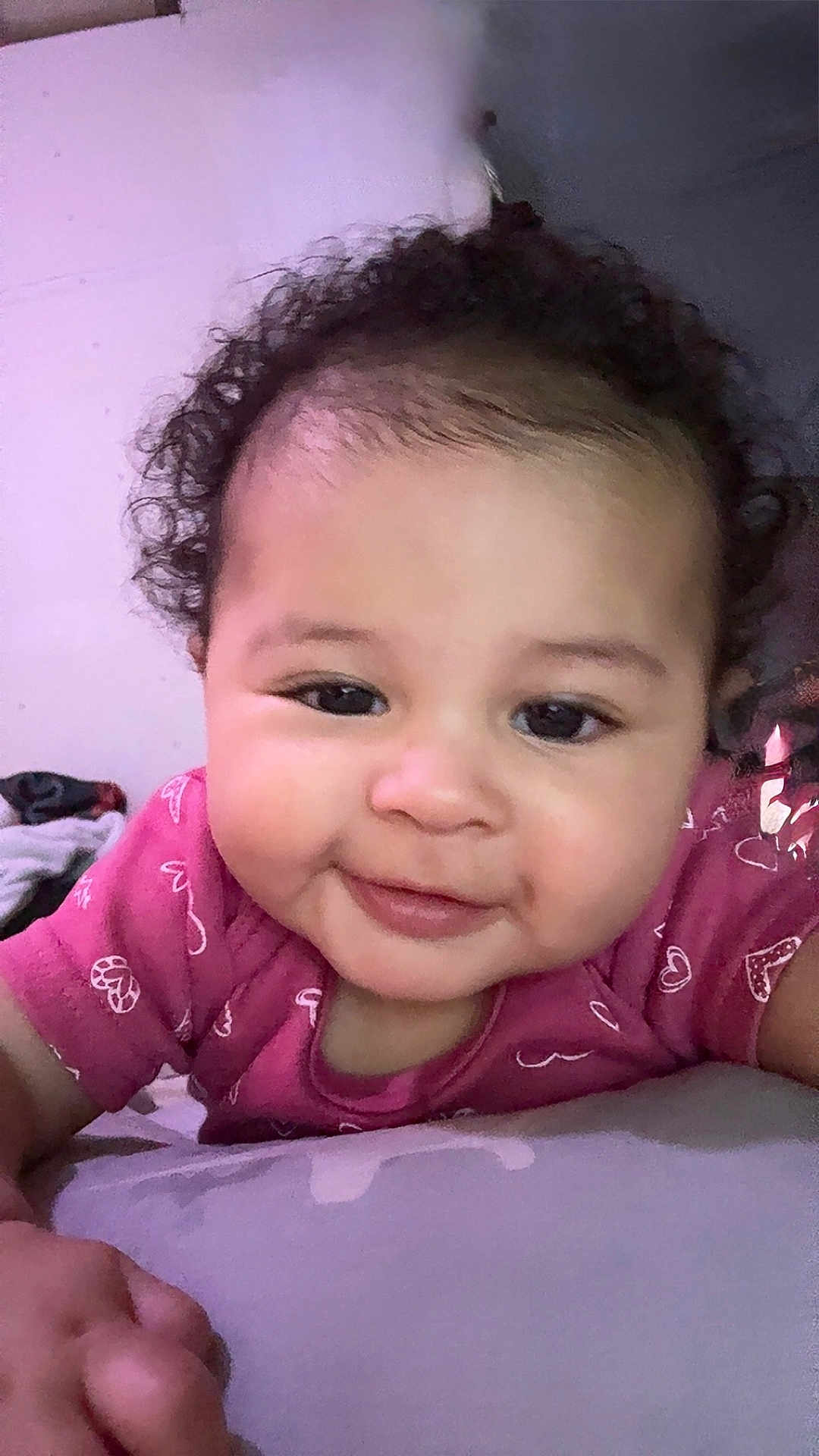 Izabella is registered to the contest to win money with this photo: baby, child, face, smile, curly_hair, pink_shirt, indoor, portrait, closeup, skin, person, infant, cute, young, lying_down, hand, soft_surface, happy, expression, head