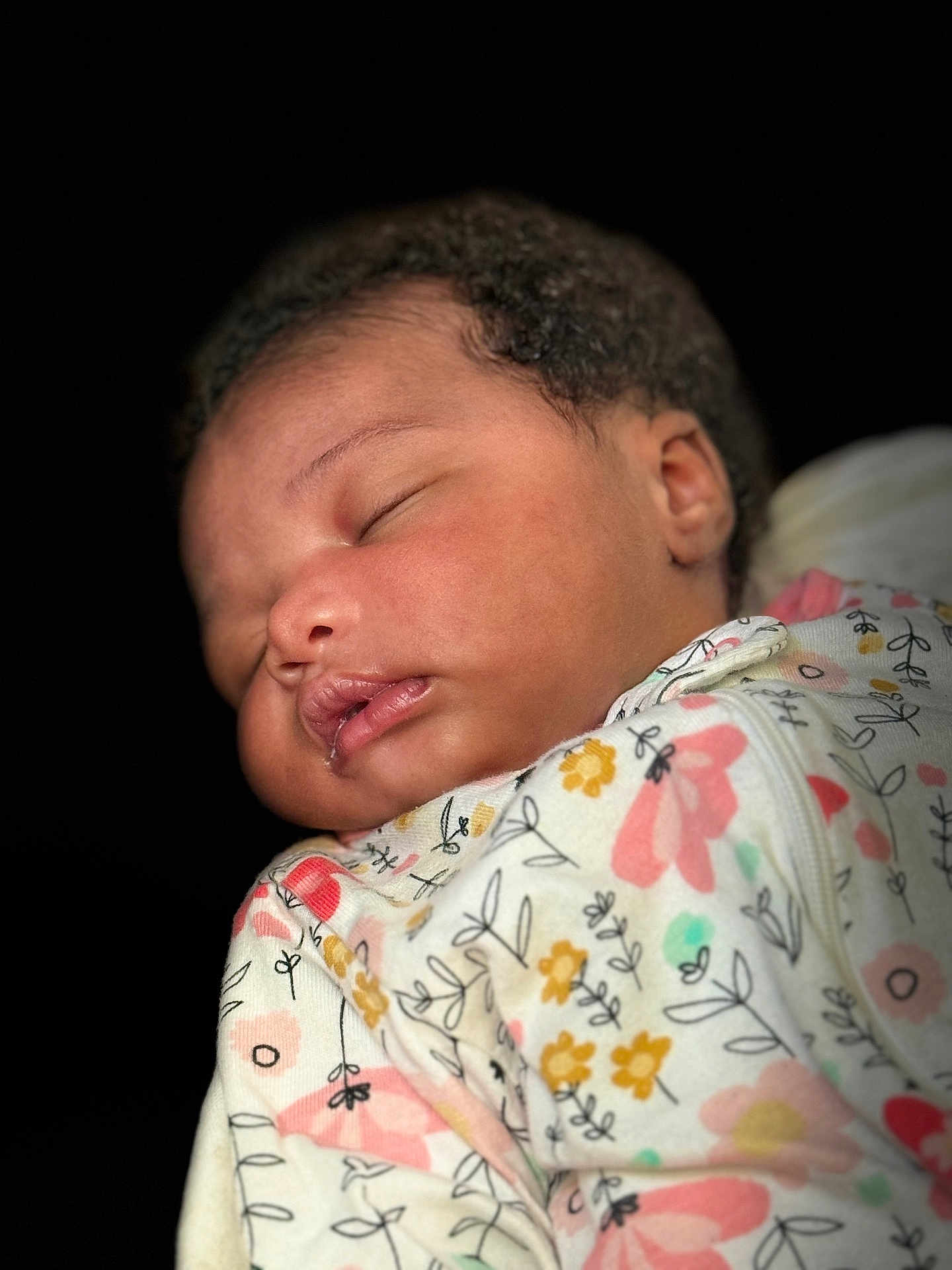 Seven is registered to the contest to win money with this photo: baby, sleeping, infant, face, curly_hair, onesie, floral_pattern, closeup, portrait, peaceful, child, soft_skin, cute, newborn, rest, cozy, human, head, expression, dark_background