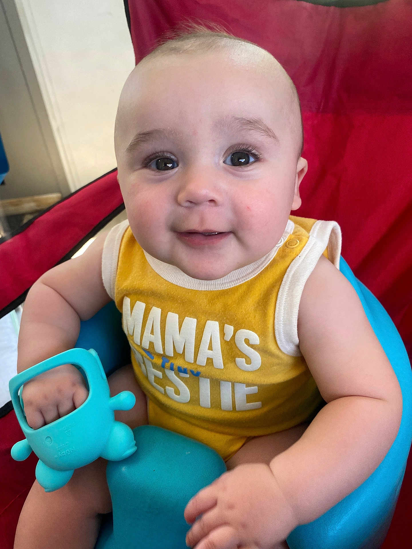 Hudson is registered to the contest to win money with this photo: baby, infant, child, smile, face, eyes, toy, blue_toy, yellow_clothing, sitting, baby_seat, red_background, arm, hand, cute, portrait, indoor, closeup, happy, skin