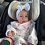 baby, infant, car_seat, headband, bow, white_clothing, floral_pattern, dress, green_socks, tongue_out, child, person, car_interior, steering_wheel, adult, seat, seatbelt, cute, smiling, closeup