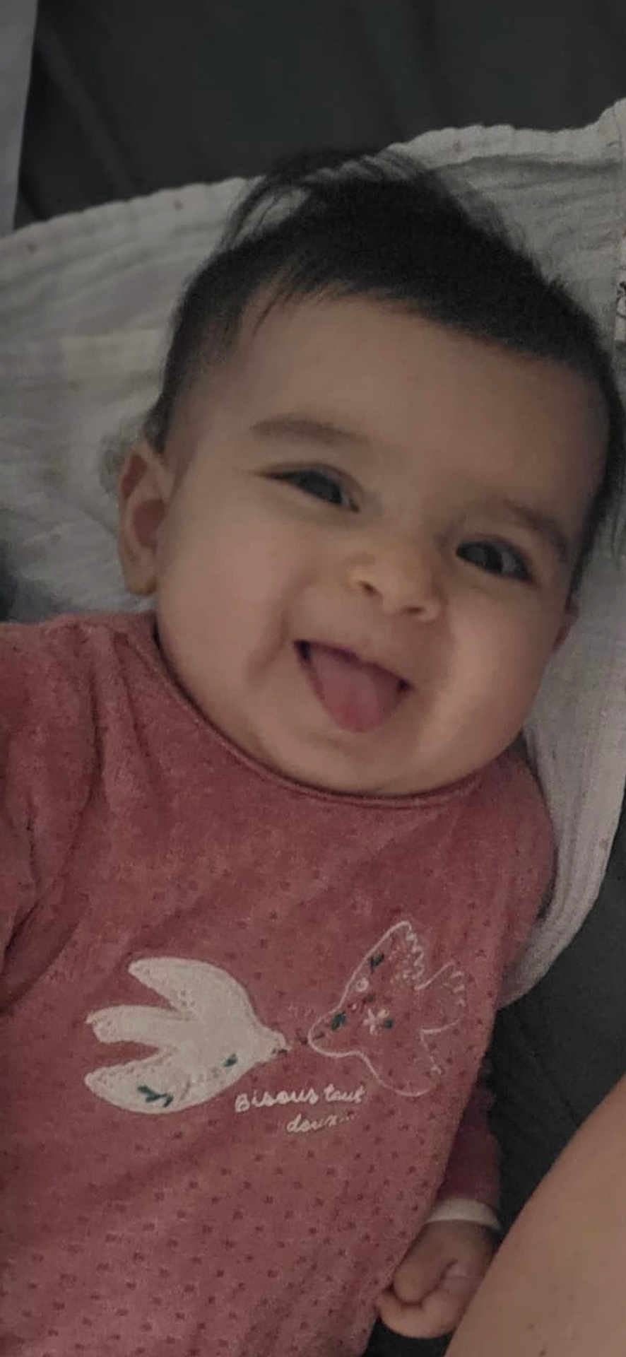 Giulia participe au concours pour gagner de l'argent avec cette photo : baby, infant, smiling, tongue_out, face, portrait, close_up, onesie, pink_clothing, bedding, blanket, chubby_cheeks, dark_hair, hand, ear, lying_down, cute, happy, bedroom, soft_lighting