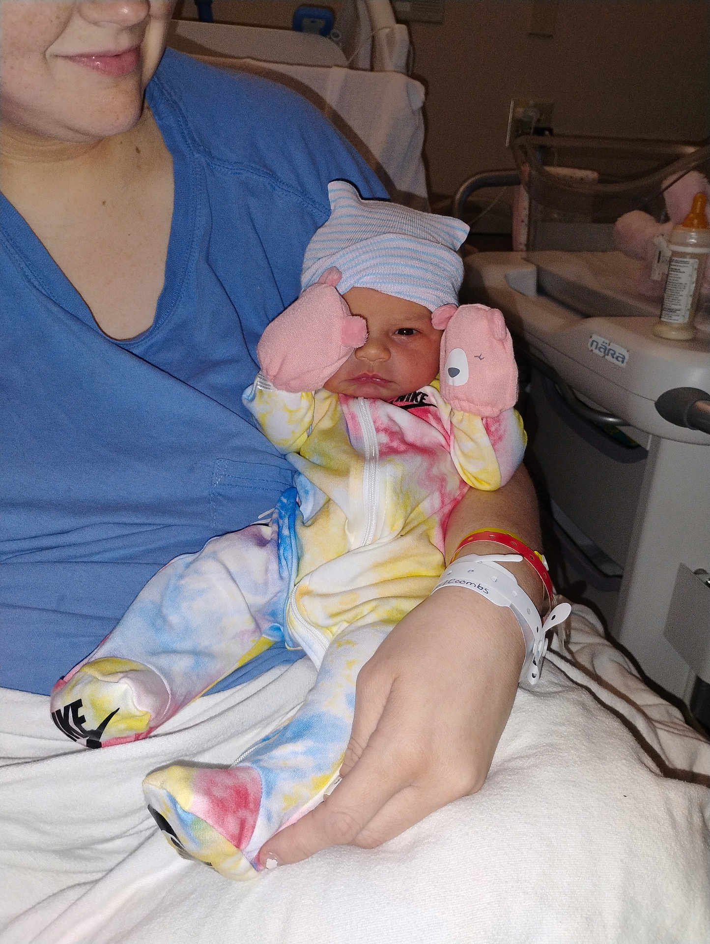 Emersyn is registered to the contest to win money with this photo: newborn, baby, infant, hospital, adult, blue_shirt, tie_dye_clothing, mittens, hospital_wristband, baby_bottle, hospital_bed, cradling, person, human_hand, face, newborn_hat, medical_equipment, bed, indoor, parent