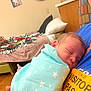 baby, newborn, swaddled, sleeping, blanket, stars, visitor_pass, blue_shirt, person, hand, bed, pillow, room, wood_floor, wall_art, bottle, blanket_tassels, furniture, indoor, peaceful
