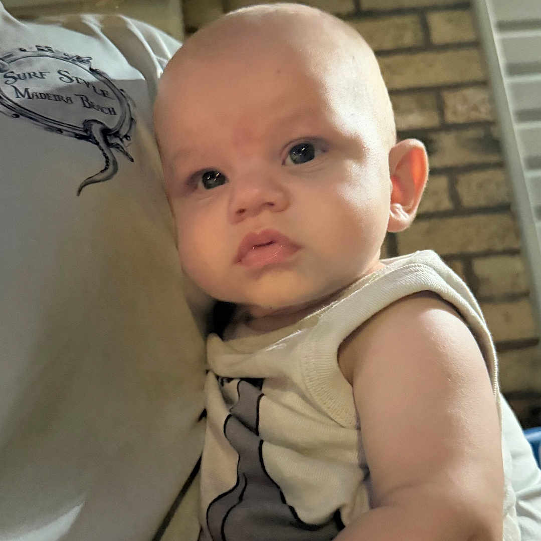 Julian is registered to the contest to win money with this photo: arm, baby, brick_wall, casual_clothing, ceiling, child, close_up, curly_hair, expression, face, glasses, holding, indoor, infant, looking, person, portrait, skin, sleeveless_shirt, young_child